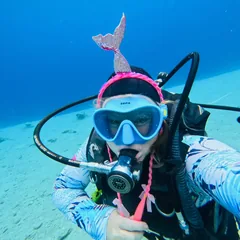 אנג׳ל מתחת למים בציוד צלילה, עם מסכה ורודה, מעל קרקעית חול.