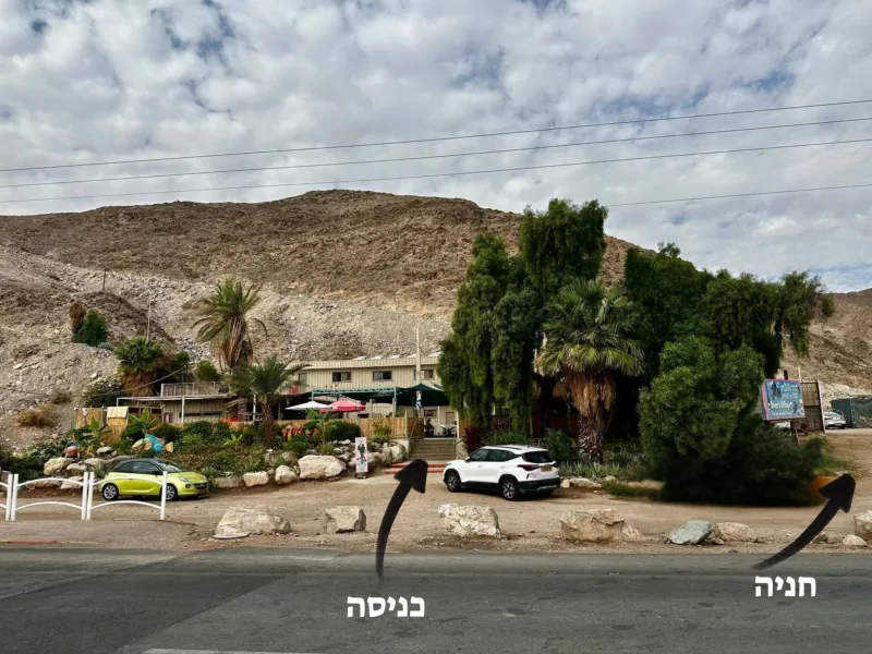מבט מהכביש אל הכפר: עצים, דקלים, מדרגות ומכוניות חונות, עם חיצים שמסומנים ״כניסה״ ו״חניה״; מאחור הר.